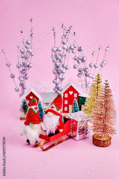 Fototapeta bright card with two gnomes in a toy forest with Christmas trees, who came home with gifts on a sled to celebrate Christmas on a pink background, vertical