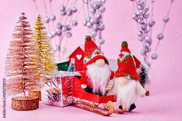Fototapeta beautiful postcard with two gnomes in a toy forest with Christmas trees, who are going to ride a sled to celebrate Christmas on a pink background, horizontal