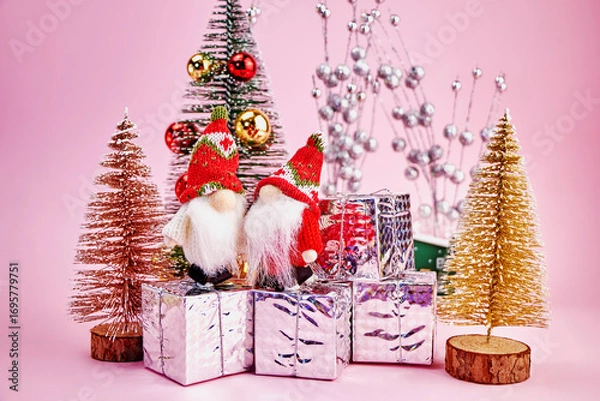 Fototapeta Christmas card on which two cute gnomes received gifts in a small forest of toy Christmas trees on a pink background, horizontal