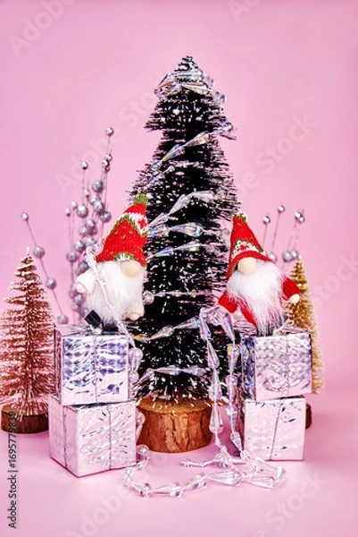 Fototapeta funny Christmas picture in which two gnomes, stand on gift boxes and decorate the tree with a silver garland on a pink background, vertically