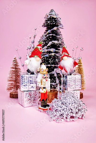 Fototapeta beautiful Christmas picture in which gnomes, standing on gift boxes and a nutcracker decorate a Christmas tree with a silver garland on a pink background, vertical
