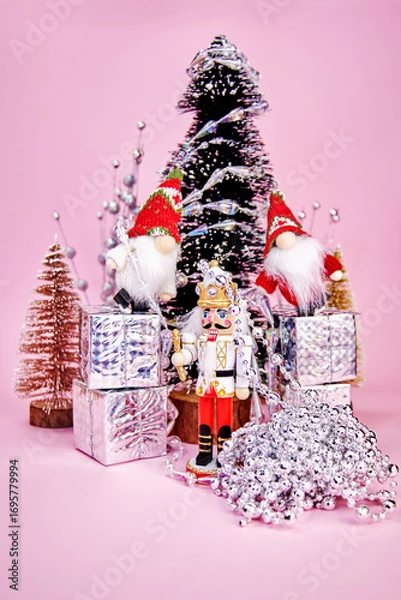 Fototapeta beautiful Christmas card on which gnomes, standing on gift boxes and a nutcracker decorate a Christmas tree with a silver garland on a pink background, vertical