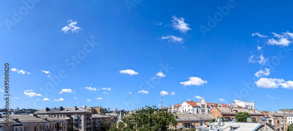 Fototapeta Panorama of isolated small clouds hanging over ordinary city buildings. Randomly scattered cloud on blue background. Clear fine summer day in capital.