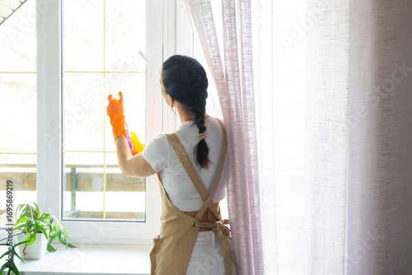 Fototapeta Woman in rubber gloves and apron manually washes window balcony of house with rag cleaner and mop inside interior. Restoring order and cleanliness in spring, cleaning servise