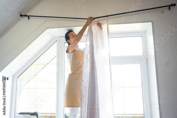Fototapeta Woman in apron hangs transparent tulle curtains on large attic windows in the house inside the white interior. Spring cleaning, tidying up