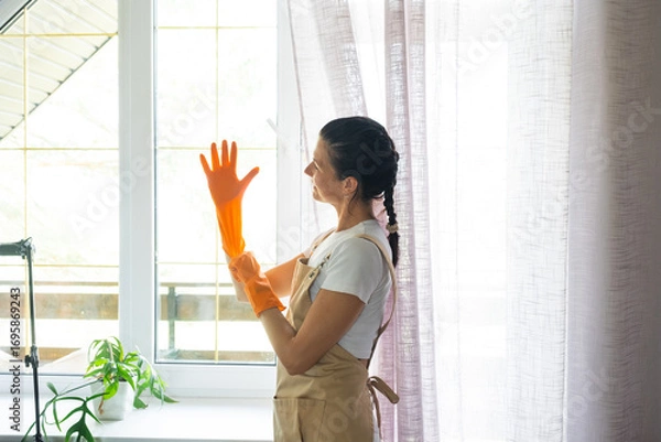 Fototapeta Woman in rubber gloves and apron manually washes window balcony of house with rag cleaner and mop inside interior. Restoring order and cleanliness in spring, cleaning servise