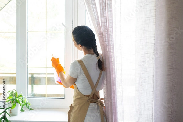 Fototapeta Woman in rubber gloves and apron manually washes window balcony of house with rag cleaner and mop inside interior. Restoring order and cleanliness in spring, cleaning servise