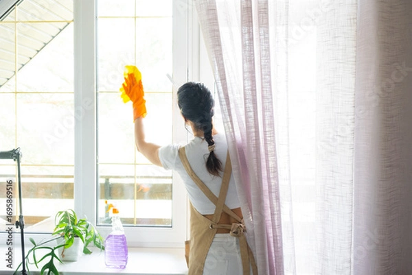 Fototapeta Woman in rubber gloves and apron manually washes window balcony of house with rag cleaner and mop inside interior. Restoring order and cleanliness in spring, cleaning servise