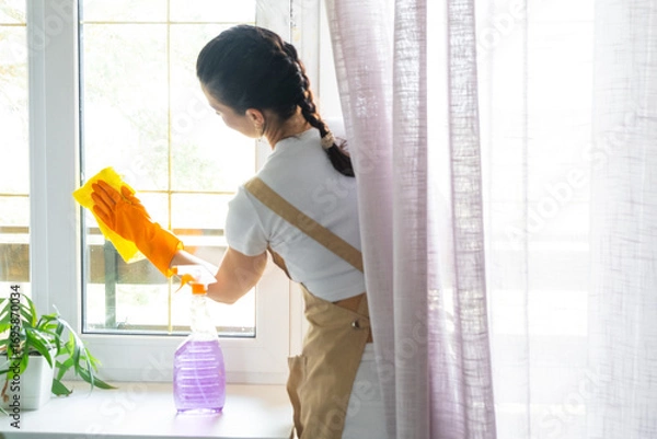 Fototapeta Woman in rubber gloves and apron manually washes window balcony of house with rag cleaner and mop inside interior. Restoring order and cleanliness in spring, cleaning servise