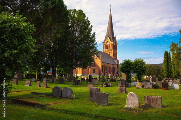 Fototapeta Brick church with tall steeple surrounded by a green cemetery and trees in Lillehammer, Norway. It is also known as Lillehammer kirke.