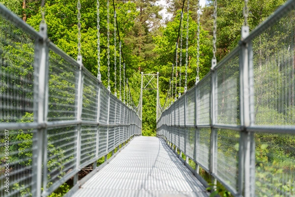 Fototapeta A long and narrow suspension bridge, supported by strong chains, winds its way through vibrant green foliage, Sweden, Knared