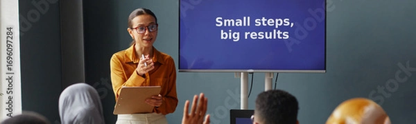 Fototapeta Website header shot of young adult Caucasian woman standing in front of interactive screen holding clipboard, teaching diverse group of students in language school classroom, students raising hands