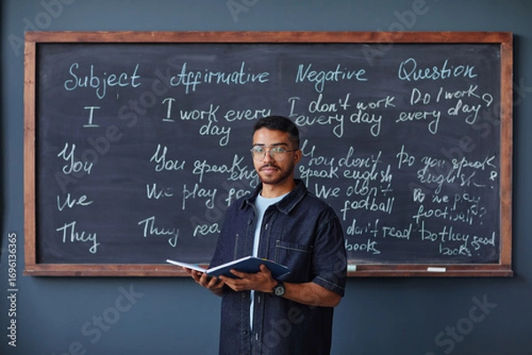 Fototapeta Portrait of young adult Black man standing in front of chalkboard holding open book, teaching English grammar lesson in language school classroom, looking directly at camera