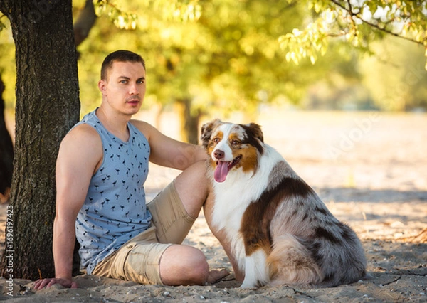 Fototapeta Photo of athletic looking slavic man with his obedient red merle australian shepherd dog sitting in front of him under the tree in the sand