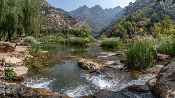 Fototapeta Mountain stream flows through a rocky landscape with lush greenery, under a clear blue sky