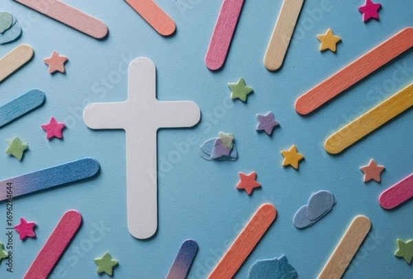 Fototapeta A centered white cross surrounded by colorful craft sticks & stars against a soft, solid blue background. Flat lay, top-down perspective