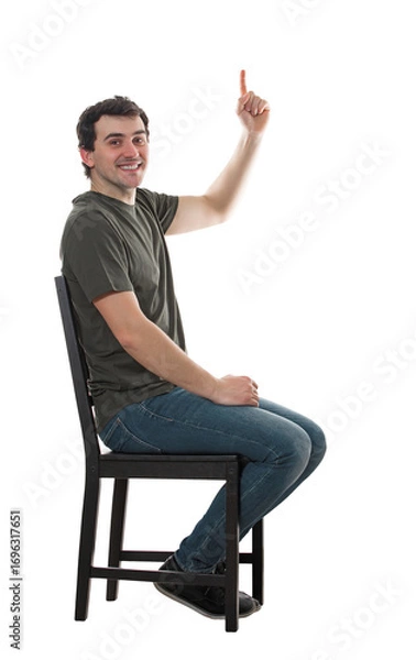 Fototapeta Side view casual guy seated on a chair pointing up forefinger, looking contented to camera. Happy man full length portrait isolated on transparent background