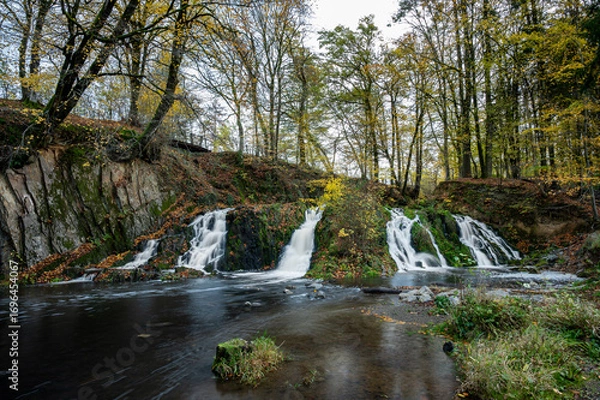 Fototapeta Long exposure captures a waterfall flowing smoothly through an autumn forest near Hirson, France, surrounded by colorful fall foliage.
