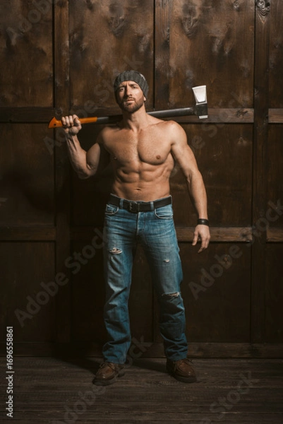 Fototapeta In a rustic setting, a muscular man stands confidently against a weathered wooden wall, holding an ax.