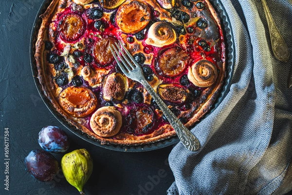 Fototapeta A tart or open pie with blueberries, figs, prunes and apricots in a baking dish on a gray background with a fork and napkin on the table. Top view. High quality photo