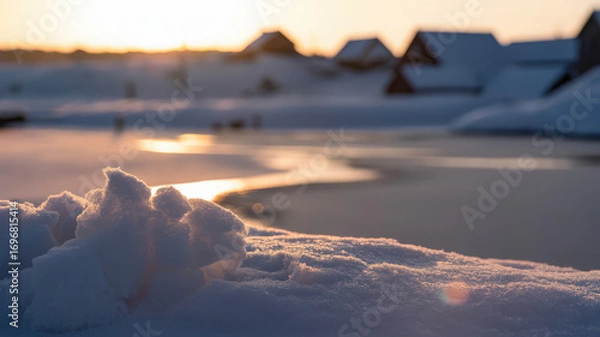 Fototapeta Snowy landscape with frozen lake and distant buildings at sunset creating a peaceful winter scene