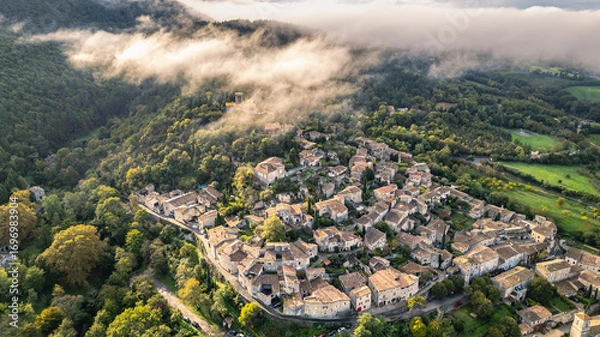 Fototapeta Aerial view of Mirmande village in France with morning fog over hills – picturesque medieval town surrounded by lush green forest and countryside landscape, historic architecture under soft misty sky