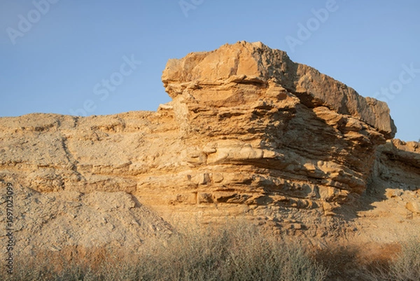 Fototapeta layered rock formation of ochre-colored stone rises from the Negev desert landscape. The eroded geological structure is set against a clear sky with sparse vegetation. Israel south