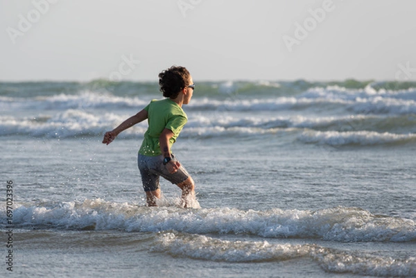 Obraz Caucasian child playing on a beach at sunset, runing through the waves and shouting towards the ocean, arms outstretched.
