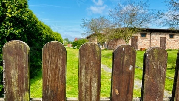 Fototapeta weathered wooden fence frames a view of a green lawn and a brick building under a bright blue sky with scattered clouds.