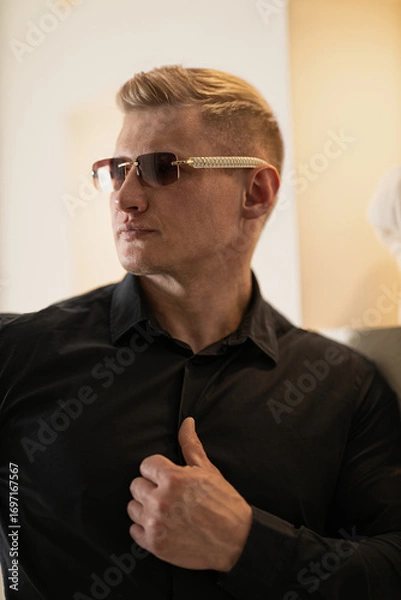 Fototapeta man wearing a black shirt and stylish glasses stands confidently showcasing his thoughtful expression. Soft warm lighting enhances the atmosphere creating a captivating indoor setting.