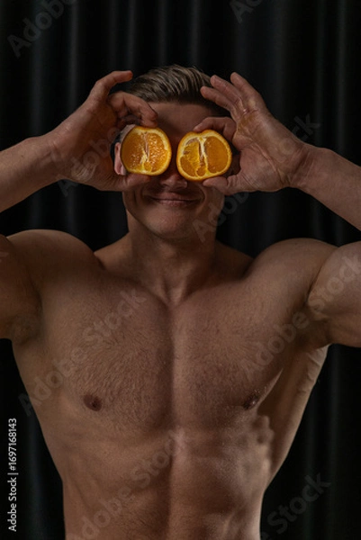 Fototapeta confident male figure with a muscular build holds two orange halves in front of his eyes smiling playfully. The dark background highlights his expression and physique.