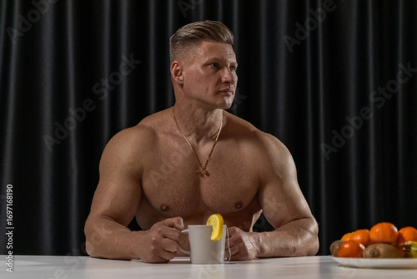 Fototapeta A muscular man sits at a kitchen table holding a mug of tea with a lemon slice showcasing his bare torso. The kitchen features dark curtains and a bowl of fruits nearby.