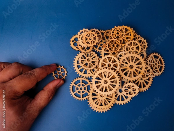 Fototapeta The image shows a hand placing a wooden gear, which is part of a larger arrangement of gears on a blue background