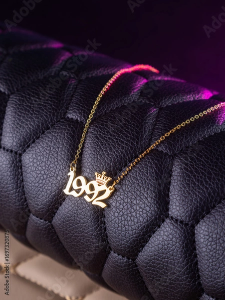 Fototapeta Close-up of a metallic gold pendant in the shape of "1992" on a chain, on a black leather background