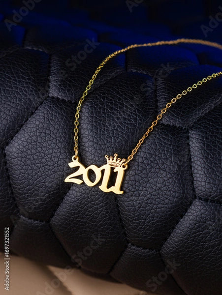 Fototapeta Close-up of a metallic gold pendant in the shape of "2011" on a chain, on a black leather background