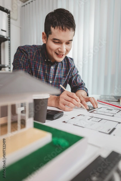 Fototapeta Young male architect working at office desk, drawing construction plans and using drafting tools, focused on building design while showing creativity and dedication in architecture.