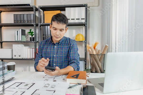 Fototapeta A young architect works on house design plans at his desk, focusing on drawings and scale models. The scene conveys creativity, dedication, and precision in modern architecture projects.