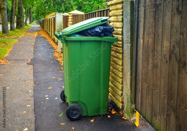 Fototapeta Green outdoor garbage bin with overflowing trash bag standing on a sidewalk near a wooden fence.
