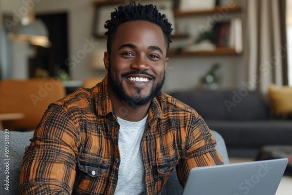 Obraz Portrait of a happy young African American man sitting on the couch at home, using a laptop, smiling at the camera while celebrating his success with a victory gesture, Generative AI