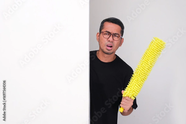 Fototapeta Adult asian man peeking behind wall holding duster with lazy face expression