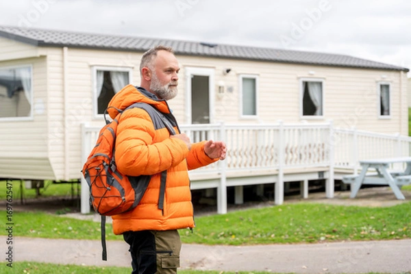 Fototapeta Middle-aged man in bright orange jacket and backpack walking near a mobile home at a campsite during an overcast day