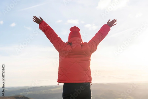 Fototapeta Person wearing a red jacket raises arms in celebration on a mountain during sunset with a vibrant sky and rolling hills in the background