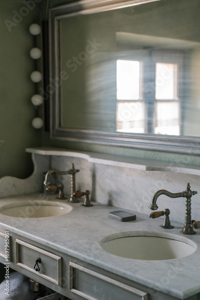 Fototapeta Vintage double sink bathroom vanity with marble countertop, antique brass faucets, and a large wall mirror. Soft natural light creates a calm and rustic atmosphere.