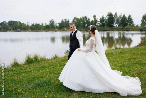 Fototapeta Wedding couple walking by serene lake during cloudy day in nature setting