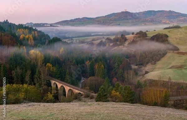 Fototapeta Pink‑tinged dawn light glows over rolling hills and fields of Banská Belá, with low fog drifting through wooded valleys, an old viaduct and autumn colors adding charm to the serene rural scene.