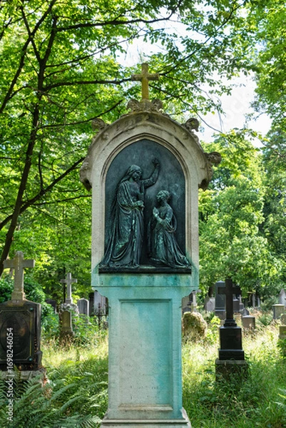 Fototapeta sehr schöner alter Grabstein mit einer Metallplatte und Bibelszene inmitten von Sträuchern Alter Südfriedhof München