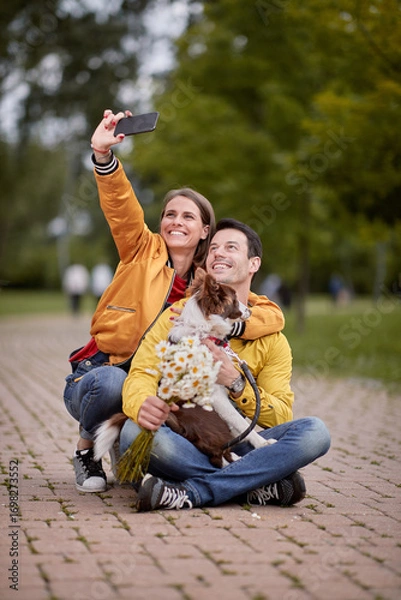 Fototapeta caucasian woman taking selfie with her boyfriend and his dog in a lap, sitting on a path in park