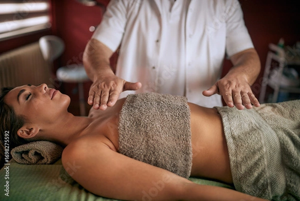 Fototapeta Young woman laying on her back, feeling relaxed and enjoying reiki massage done by a male therapist in a modern studio.