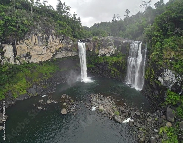 Fototapeta Two waterfalls cascading into a dark pool, surrounded by lush green forest and rocky cliffs