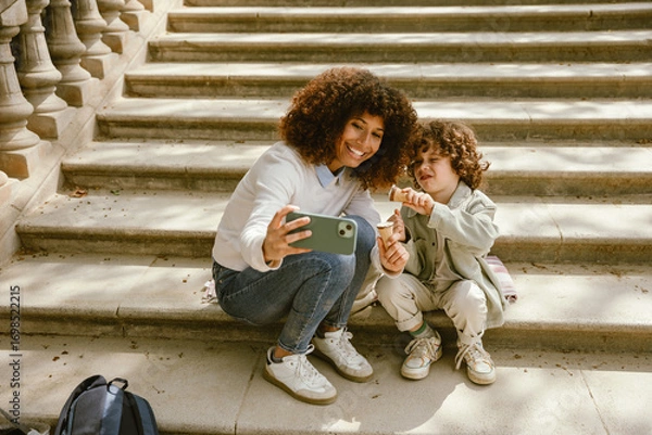 Fototapeta Mother and son holding ice cream and posing while sitting on a blanket on the stairs and looking at the phone she is holding
